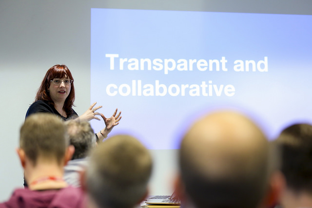 Photo of a woman giving a talk in front of an audience. Slide on the screen reads "Transparent and collaborative". Audience look engaged, and speaker is gesticulating.