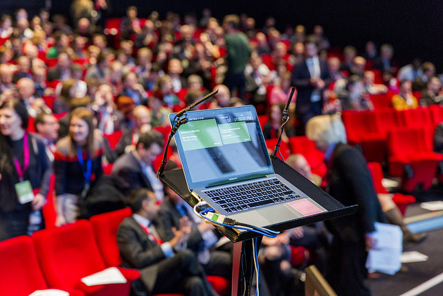 Crowd of people in an auditorium (taken at Sprint 16 event) with laptop in the foreground with a presentation loaded onto it.