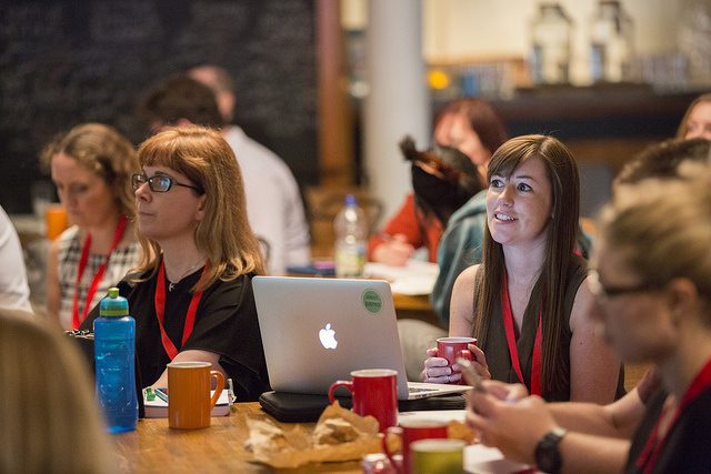 Photo from #GovBlogCamp - audience members sat around tables intently watching the action onstage. Warm, relaxed atmosphere, with macbooks and coffee cups on the tables.