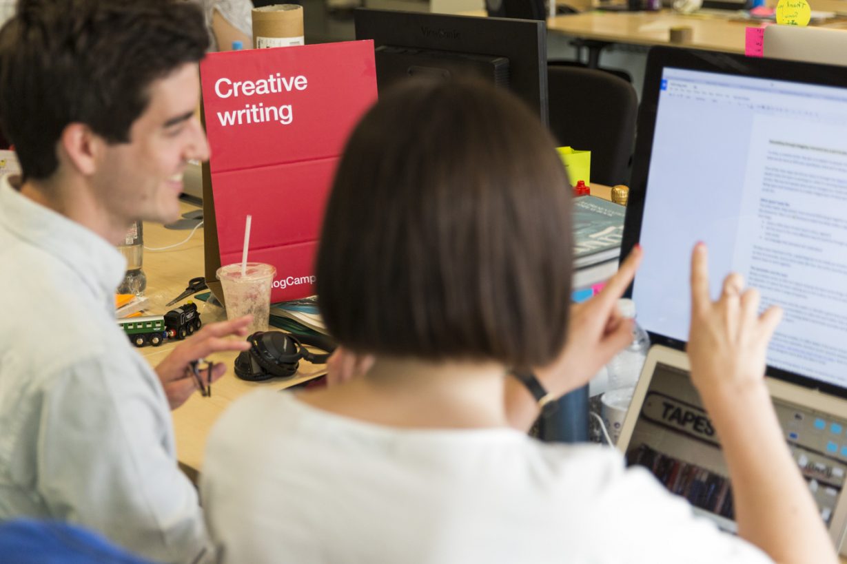 Amy McNichol and colleague sitting at a computer writing a piece of content together. Featuring a sign on the table that reads 'creative writing'.