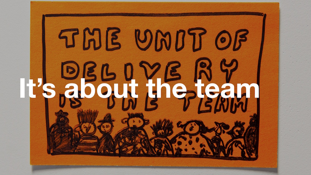 Post-it note showing a Paul Downey illustration of people with the caption 'The unit of delivery is the team' - with 'It's about the team' superimposed over the top