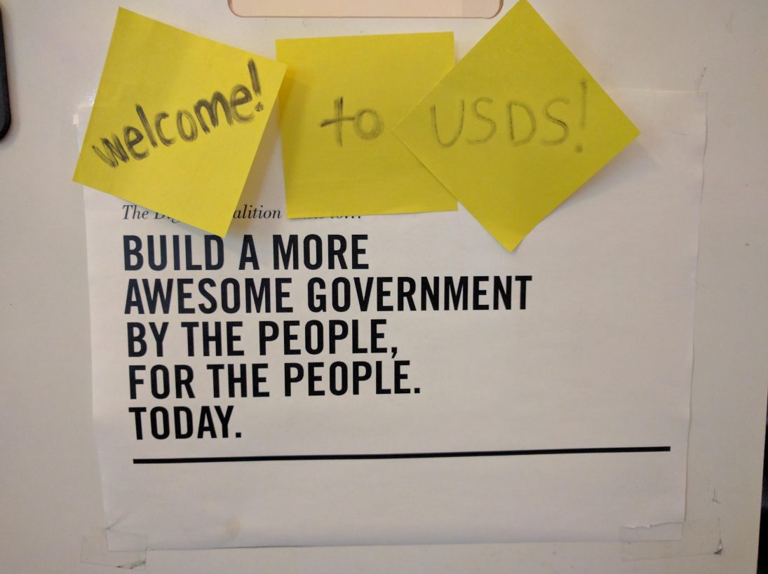 A sign that says 'Build a more awesome government by the people, for the people. Today.' The sign has sticky notes on it that say 'Welcome! to USDS!'