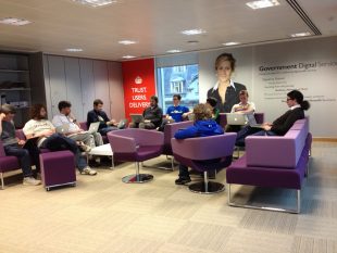 10 members of the GDS team sit with their laptops in the seating area on the 6th floor of Aviation House. There is a large wall with a picture of Martha Lane Fox and a poster saying ‘TRUST. USERS. DELIVERY’ on the wall behind them.