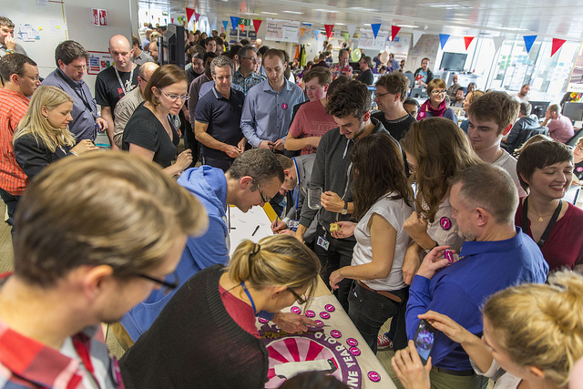 Over 100 members of the GDS team crowd around a table to get cake and pin badges to celebrate the 1st year of GOV.UK