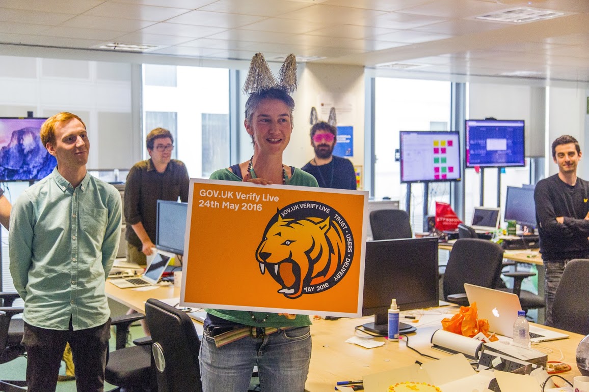 Photo of the the GOV.UK Verify team celebrating going live. Team leader Janet Hughes is shown holding a sign saying “GOV.UK Verify Live. 24 May 2016”.