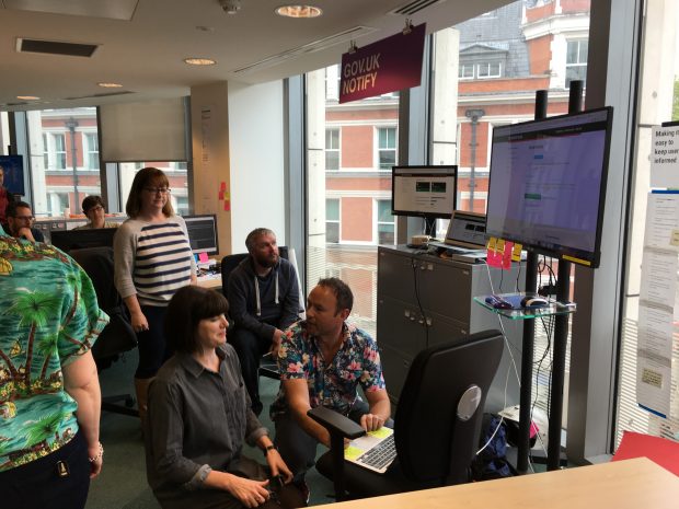 Photo showing the GOV.UK Notify team gathered around a computer and a large external monitor to send the first messages via the service.