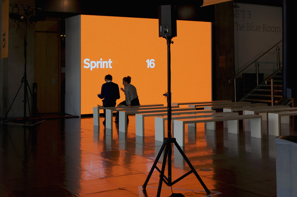 Photo of Sprint 16 venue, taken shortly before the doors opened. Two members of the production team sit in front of a large screen showing the “Sprint 16” branding on a bright orange background.