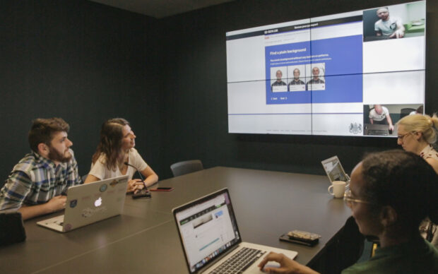 user research lab at GDS with a group of 4 people sitting at a table with their laptops and a bit screen showing user research participants in the background