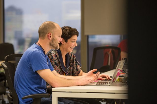 two people sitting at a desk together with laptops