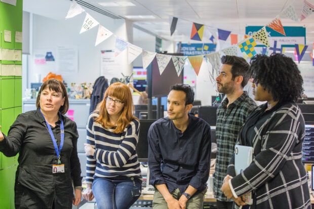 GOV.UK team standup, showing 5 members of the team looking at a board and one of them talking to the rest