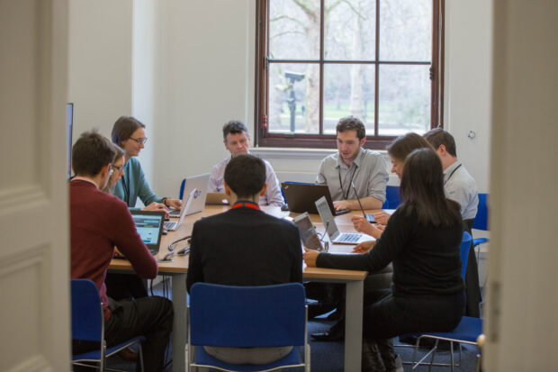 9 people sitting around a table, looking at their laptops