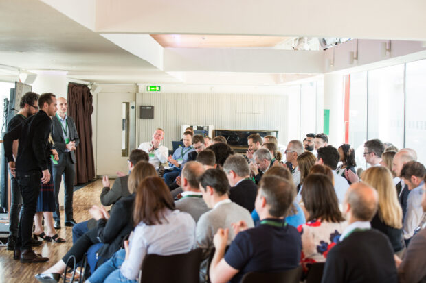 A room full of people listening to 3 presenters standing on the stage