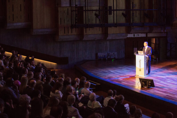 John Manzoni speaking at Sprint 18, standing on the stage, people in the auditorium visible in the shadow