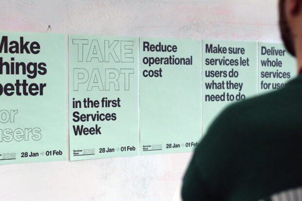 A wall with Service Week posters on it. One says "Make things better for users', a second says "Take part in the first Services Week", a third says "Reduce operational cost", a fourth says "Make sure services let users do what they need to do" and a fifth says "Deliver whole services for users"