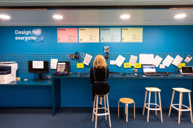 The GDS accessibility lab - a blue wall with a table with laptops and other devices on it. A user sitting at the table doing some work.