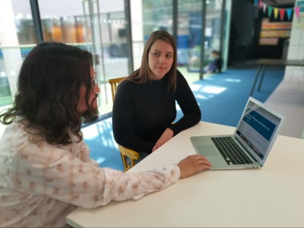 two people sitting at a table, one of them looking at a laptop, the other one facing the camera