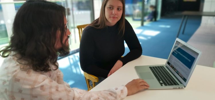 two people sitting at a table, one of them looking at a laptop, the other one facing the camera