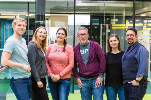 6 people standing in a row, facing the camera, in front of a window