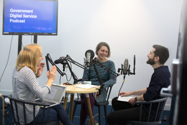 3 people sitting around a table in a recording studio, speaking to microphones. There is a screen behind them that says 'Government Digital Service Podcast'