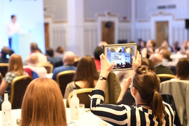 An audience facing away from the camera, one person in the foreground holding up an ipad and taking a picture of the presenter, who is visible in the background but blurry