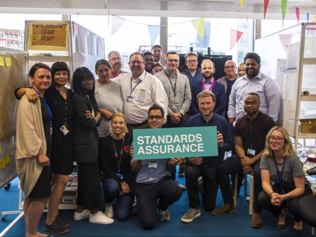 a big group of people posing for a picture, facing the camera and holding a sign that says 'standards assurance'