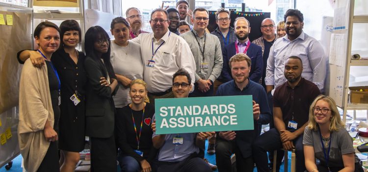 a big group of people posing for a picture, facing the camera and holding a sign that says 'standards assurance'