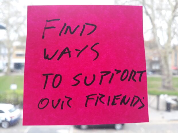 A post-it note on a window, saying 'find ways to support our friends'