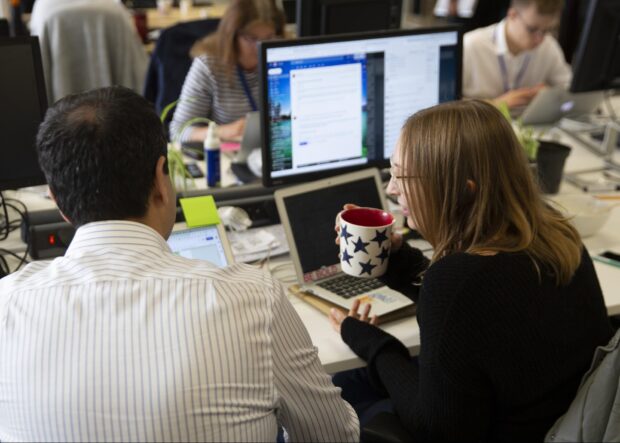 2 people sitting at a desk, looking at a laptop screen together. Other people working in the background