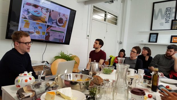 people sitting around a table, listening to a presenter and looking at a screen