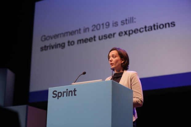 Jen Allum on the stage, with a screen in the background reading 'Government in 2019 is still striving to meet user expectations'