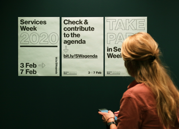 A woman holding a phone while looking at posters for Services Week 2020. She looks at one poster saying ‘Check & contribute to the agenda’, which includes an internet address to the open agenda: bit.ly/SWagenda