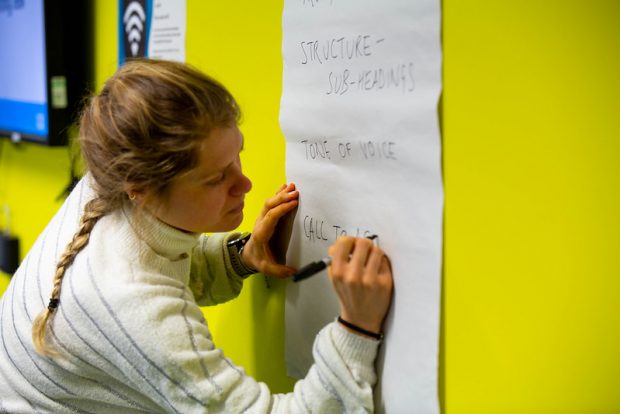 A women is writing on a piece of paper fixed on a wall. She has written the following bullet points: "Structure - subheadings", "Tone of voice", and she is beginning to write "Call to action".