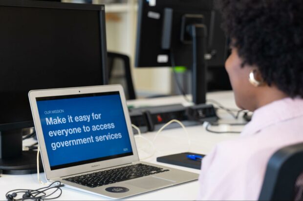 A person is looking at their laptop screen, which reads "Our Mission: Make it easy for everyone to access government services".