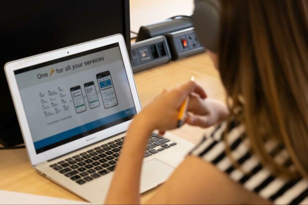 A person is looking at their laptop screen, which reads "One key for all your services", using a key emoji as a substitute. There are also visualisations of a smartphone screen below the text.