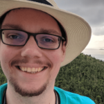 Jonathan is wearing a hat and smiling. In the background a forest and the sea with little islands and rocks are visible.
