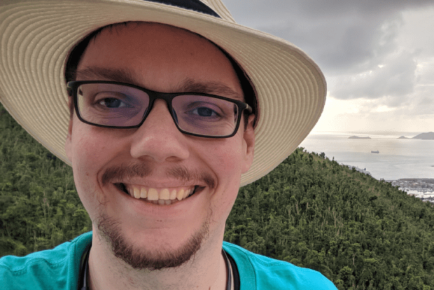 Jonathan is wearing a hat and smiling. In the background a forest and the sea with little islands and rocks are visible.