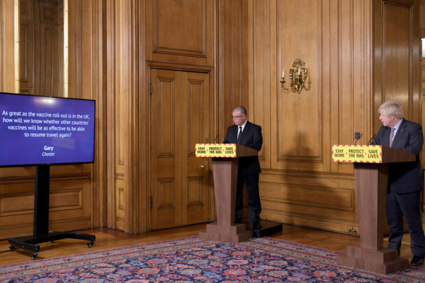 The Prime Minister, Boris Johnson, and another person are hosting a coronavirus press conference. They are looking at a TV screen displaying a question from Gary in Chester: "As great as the vaccine roll-out is in the UK, how will we know whether other countries' vaccines will be as effective to be able to resume travel again?"