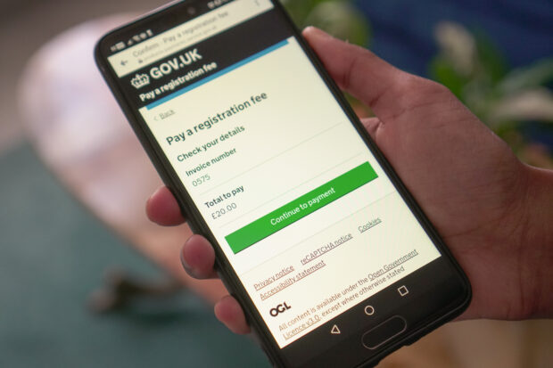 A mobile held in someone's hand. On the phone there is a GOV.UK payment page. The title of the page is 'Pay a registration fee' and the invoice and total amount to pay is shown.
