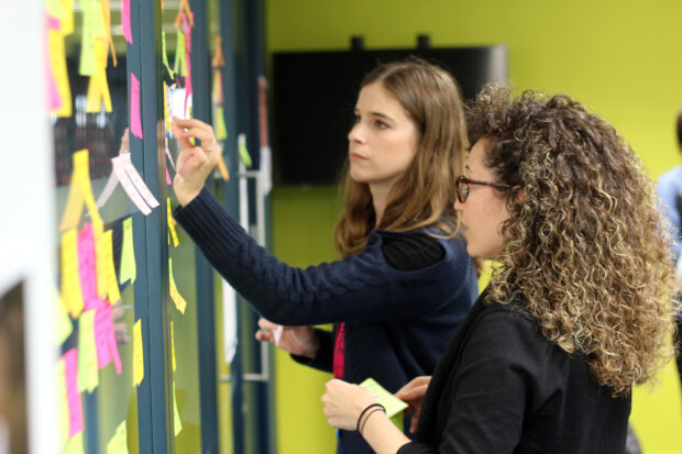 Two women working on user research with a wall of colourful post it notes