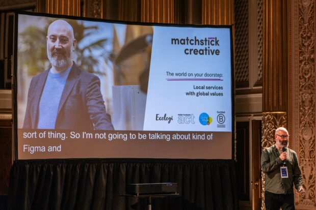 Greg MaCoy from Matchstick Creative gives his talk on stage at Design System Day. He stands on stage in front of a large screen, showing his picture and title of his talk. The world on your doorstep: Local services with global values.