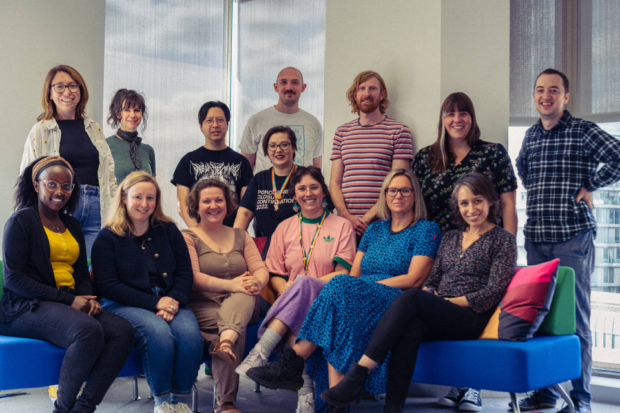 Picture of the GOV.UK Forms team with 8 members standing behind 6 members of the team seated on a blue corner sofa.