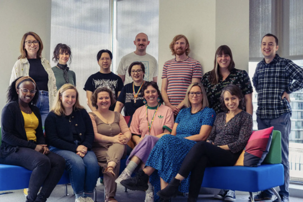 Picture of the GOV.UK Forms team with 8 members standing behind 6 members of the team seated on a blue corner sofa.