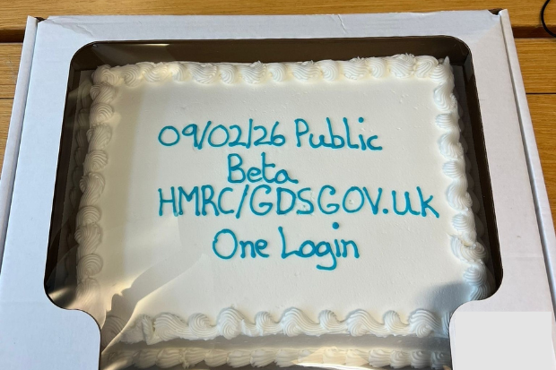 A decorated cake with white icing. "09/02/26 Public Beta HMRC/GDS GOV.UK One Login" is written on it in blue icing. 